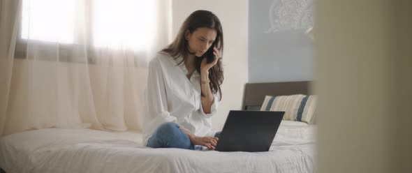 A woman talking to someone on the phone while working with a laptop from home. 