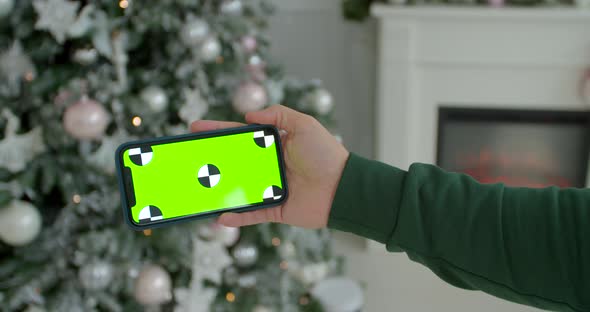 Man Holding a Mock-up Smartphone Greenscreen Chrome Key in New Year Decorated Room. Closeup