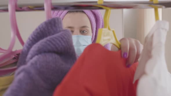 Portrait of Young Caucasian Woman in Face Mask Standing Behind Garment Rack and Choosing Outfit