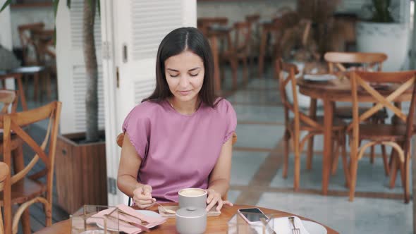 Young Woman Drinks Coffee in Cafe and Puts Sugar in Beverage, Stirs with Spoon