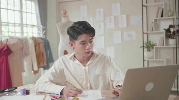 Young Asian Male Designer Working On Laptop And Sketching At Desk In Studio