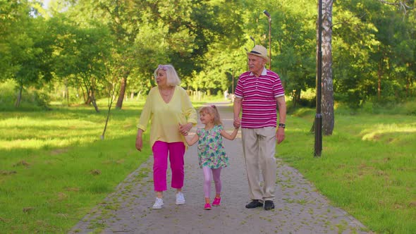 Child Girl Kid Walking with Senior Grandmother and Grandfather Family Holding Hands in Summer Park