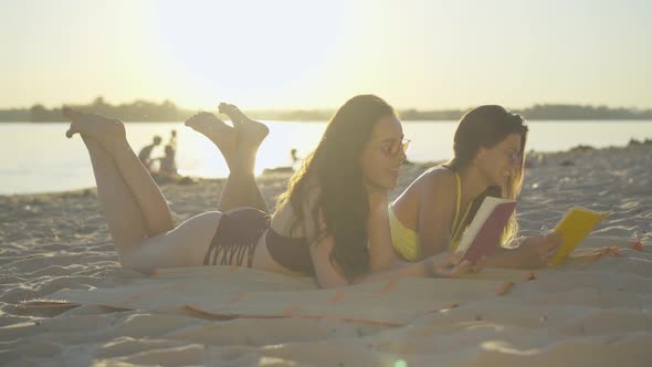 Side View Wide Shot of Relaxed Young Women Reading Books at Sunset on Sandy Beach. Portrait of