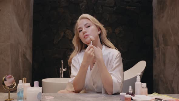 Young Blonde Woman in a White Robe in the Bathroom with a Roller Massager