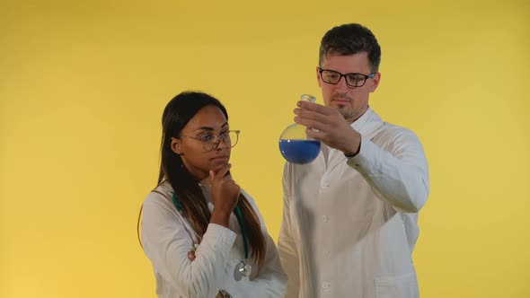 Multiethnic Man and Woman in Lab Coats Looking on Flask with Experimental Liquid on Yellow