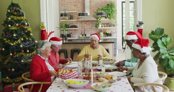 Group of happy diverse senior friends in santa hats eating christmas dinner together at home