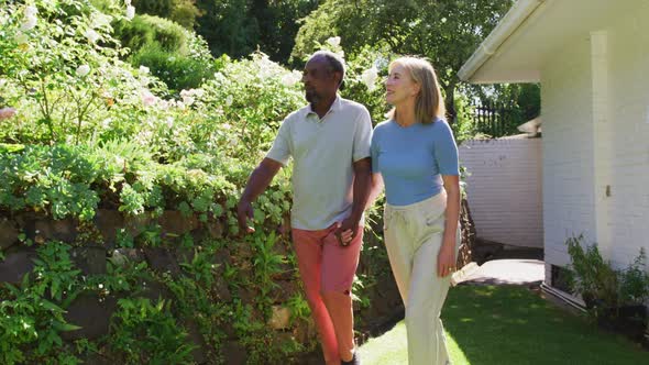 Diverse senior couple walking in their garden in the sun holding hands and talking