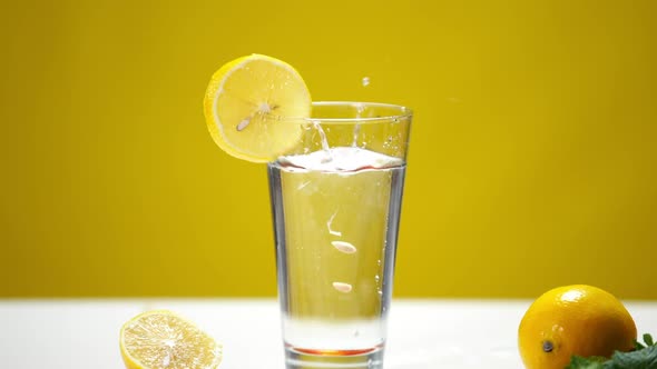 Closeup Glass with Refreshing Lemon Water with Droplets and Fruit Pits Falling Inside in Slow Motion