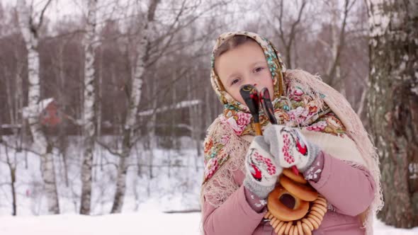 Cute girl in a traditional Russian headscarf and mittens playing on spoons on winter forest backgrou