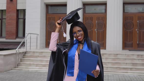 A Shy African American Female Graduate in a Black Mantle of a Master's Hat Raises Her Hat