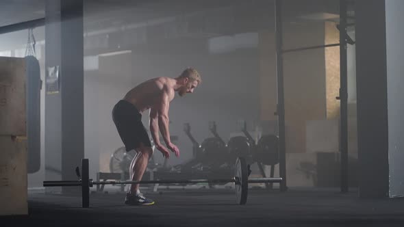 A Young Man Without a Shirt in Slow Motion Jumps a Burpee Over a Barbell in the Gym in Backlight