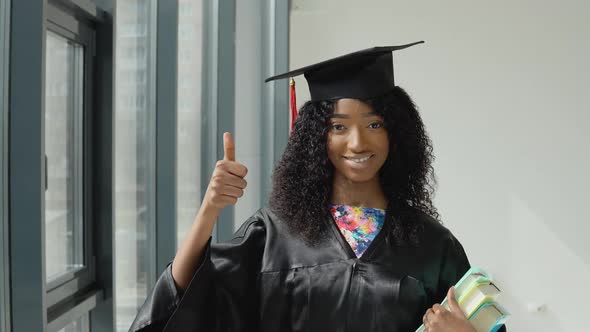 Young African American Female University Graduate Stands with Textbooks in One Hand and Shows