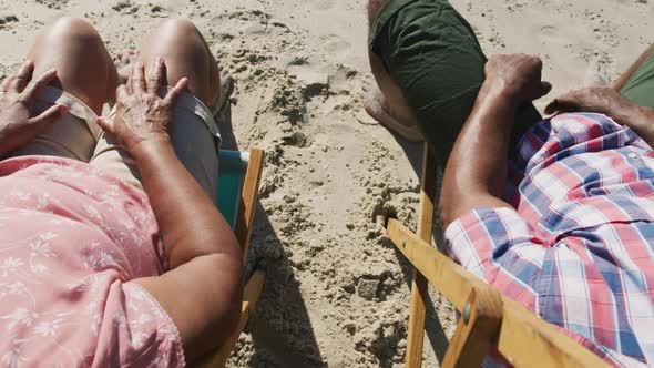 Senior african american couple sitting on sunbeds and holding hands at the beach