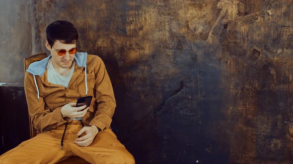 Stylish Man in Bright Glasses Sits on Chair Browsing Mobile Phone Near Shabby Brown Wall