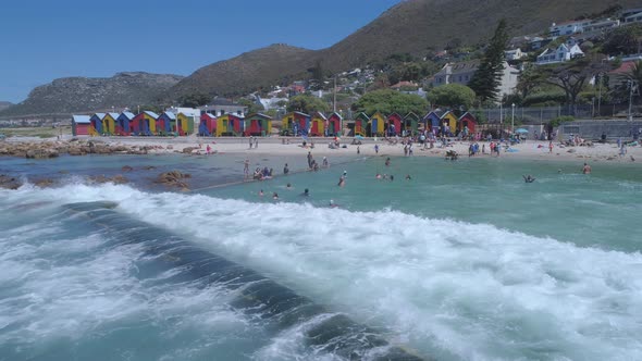 Drone hovers as wave crashes into ocean tide pool, coloured beach shacks in background