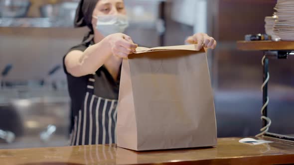 Blurred Caucasian Young Woman in Covid Face Mask Packing Takeaway Food in Cafe or Restaurant