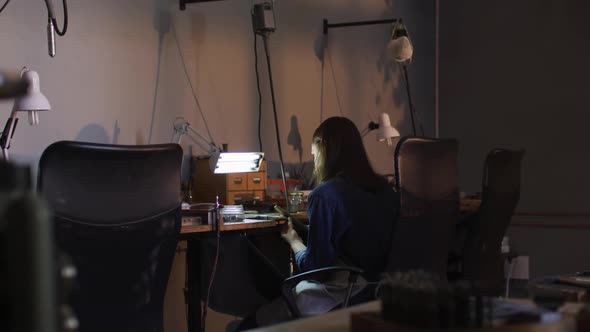 Back view of caucasian female jeweller sitting at desk, making jewelry in workshop