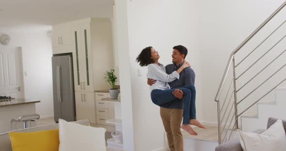 Happy romantic hispanic man carrying woman downstairs in living room