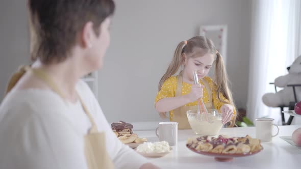 Serious Little Girl Beating Ingredients in Bowl and Smiling To Blurred Woman at the Foreground