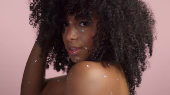 Mixed Race Black Woman with Curly Hair Covered By Crystal Makeup on Pink Background in Studio