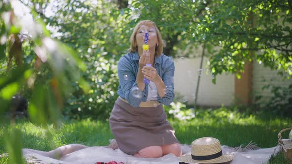 Wide Shot Portrait of Relaxed Happy Senior Woman Blowing Soap Bubbles in Slow Motion Sitting on