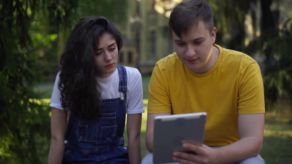 Young Intelligent Students Talking Surfing Internet on Tablet Sitting on Bench at College Yard