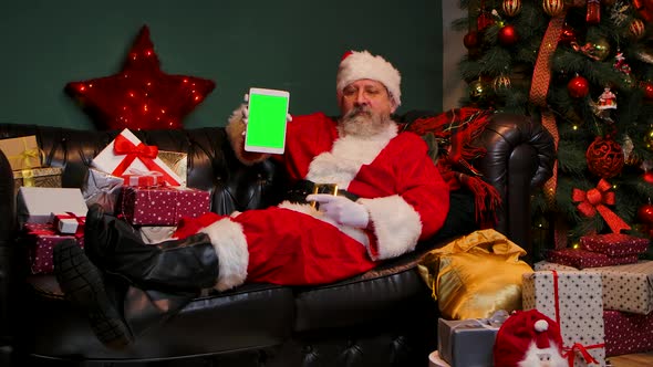 Santa Claus Lies on the Sofa Near Decorated Christmas Tree and Gift Boxes Shows Tablet with a Green