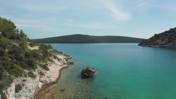 Aerial shot of a small beach with a rusted ship wreck in the foreground. Drone flies over the wreck.