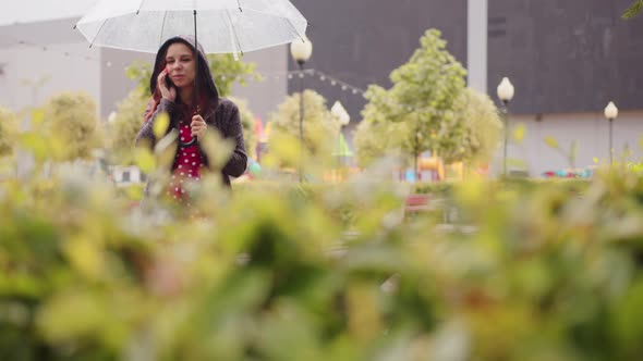 Young Beautiful Woman with Transparent Umbrella Talking on Mobile Phone in Rain on City Street