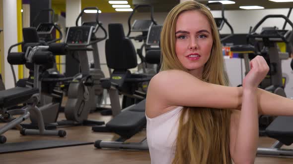 A Young Beautiful Woman Stretches Her Hands in a Gym and Smiles at the Camera - Closeup