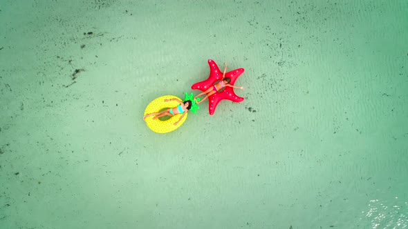 Aerial view of two girls floating on inflatable mattress in transparent sea, holding each other.