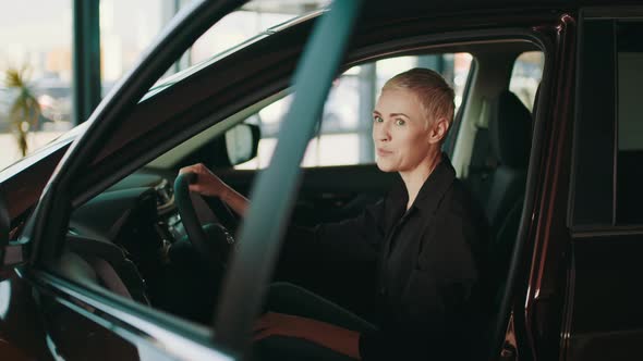 Cheerful Blond Woman Sitting in Modern Car with Open Door and Looking Aside