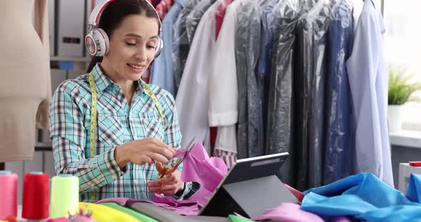 Woman Designer Stylist in Headphones Cuts Fabric at Workplace in Atelier