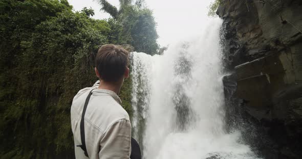 Low Angle Shot Circling Around a Young Male Explorer Pointing at the Large Waterfall in the Jungle