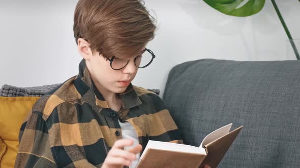 A Teenage Boy with Glasses Enthusiastically Reads a Book Sitting at Home on the Couch