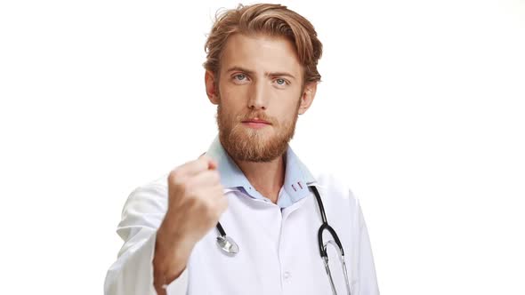 Severe Young Caucasian Male Doctor with Light Beard Wearing Medical Overall Showing Fist at Camera