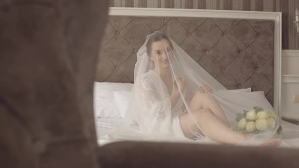 Lovely Stylish Bride Woman in Night Gown Under Veil with Bouquet Lie Down on Bed in Home Bedroom