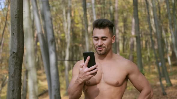 Strong Guy With Naked Torso Stands In Autumn Park And Speaks On Mobile Phone.