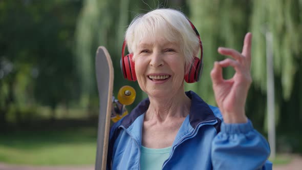 Cheerful Fit Senior Woman with Skateboard in Headphones Gesturing OK Looking at Camera Smiling