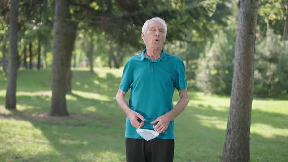 Medium Shot of Caucasian Senior Man Taking Off Coronavirus Face Mask Inhaling Fresh Air in Park