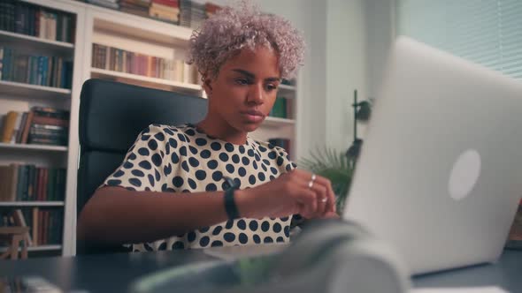 Young African American Woman Bends Over to Laptop Sits at Desk in Office
