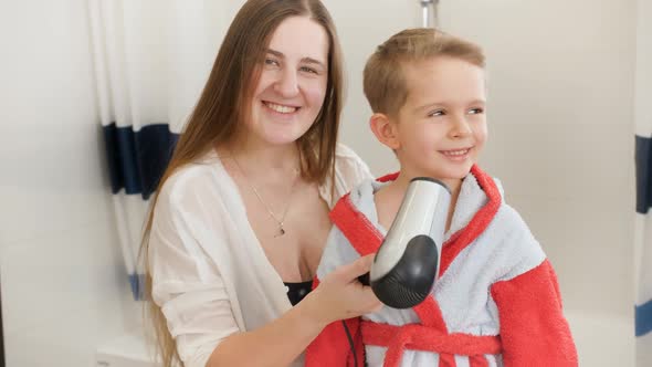 Beautiful Smiling Woman Drying Hair and Blowing Hot Air with Hairdryer on Her Little Son After