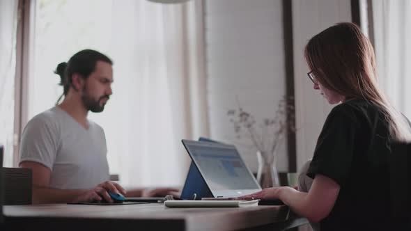 A young man and a young girl are working in front of laptops in their bright apartment.