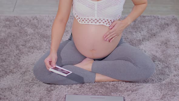 Top view Pregnant Woman sitting on carpet using computer laptop and holding ultrasound image