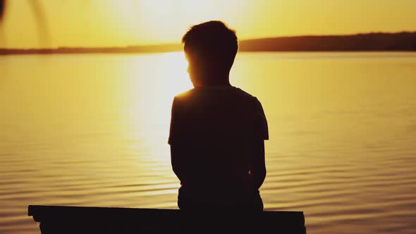 Little boy sitting on wooden dock at sunset. Child is watching the sunset