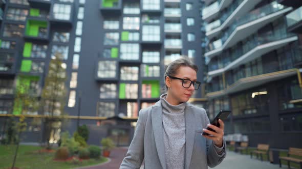 Woman Walking on a Business District Holds Takeaway Coffee Shopping Bags and Using Smartphone