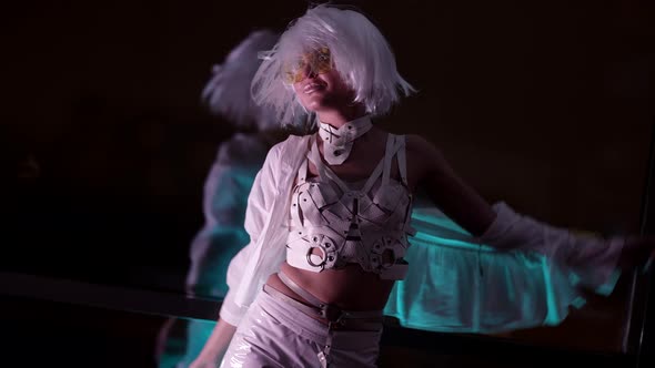 A Blonde Woman in Stylish Glasses in an Unusual White Sexy Suit Poses in the Studio Against the