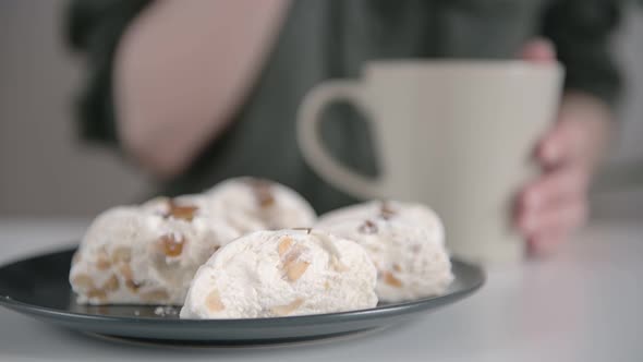 Lady in dark shirt eats large white oriental sweets slice from plate and drinks beverage from mug