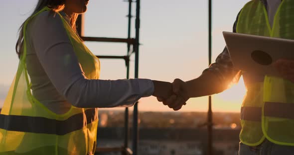 Builders Man with a Tablet and a Woman in White Helmets Shake Hands at Sunset Standing on the Roof alt