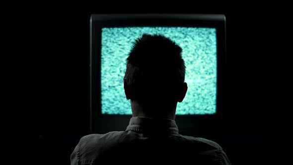 Rear View of a Silhouette of a Man Sitting in Front of a Flickering TV in a Dark Studio on a Black